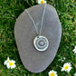 Fine silver Molly and Me Jewellery daisy disc pendant displayed on a pebble set on a background of grass with daisies