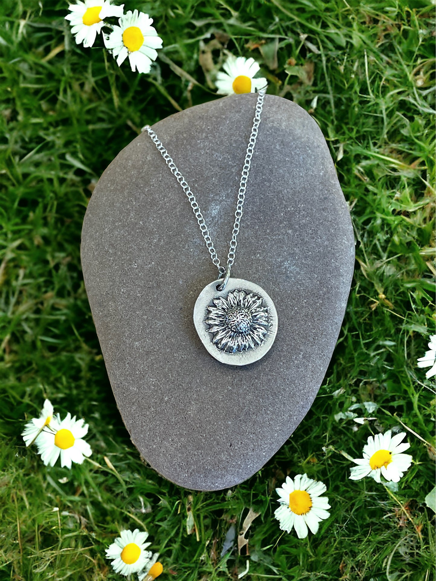 Fine silver Molly and Me Jewellery daisy disc pendant displayed on a pebble set on a background of grass with daisies