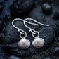 A pair of silver dangle earrings shaped like textured scallop or cockle shells, resting on dark wet rocks with small pebbles and seaweed around them.