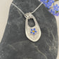 Handmade Silver pendant featuring a single hand painted forget me not flower on a textured background with a cut out design and 3 jump rings. The pendant is laying on a stone and has some forget me not flowers in the background.