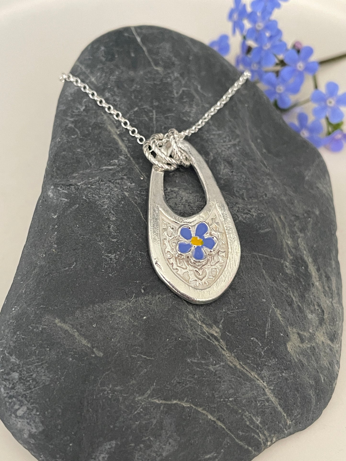 Handmade Silver pendant featuring a single hand painted forget me not flower on a textured background with a cut out design and 3 jump rings. The pendant is laying on a stone and has some forget me not flowers in the background.