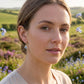 A woman, wearing small blue forget me not teardrop earrings, photographed outdoors in a field of purple flowers under a bright sky.