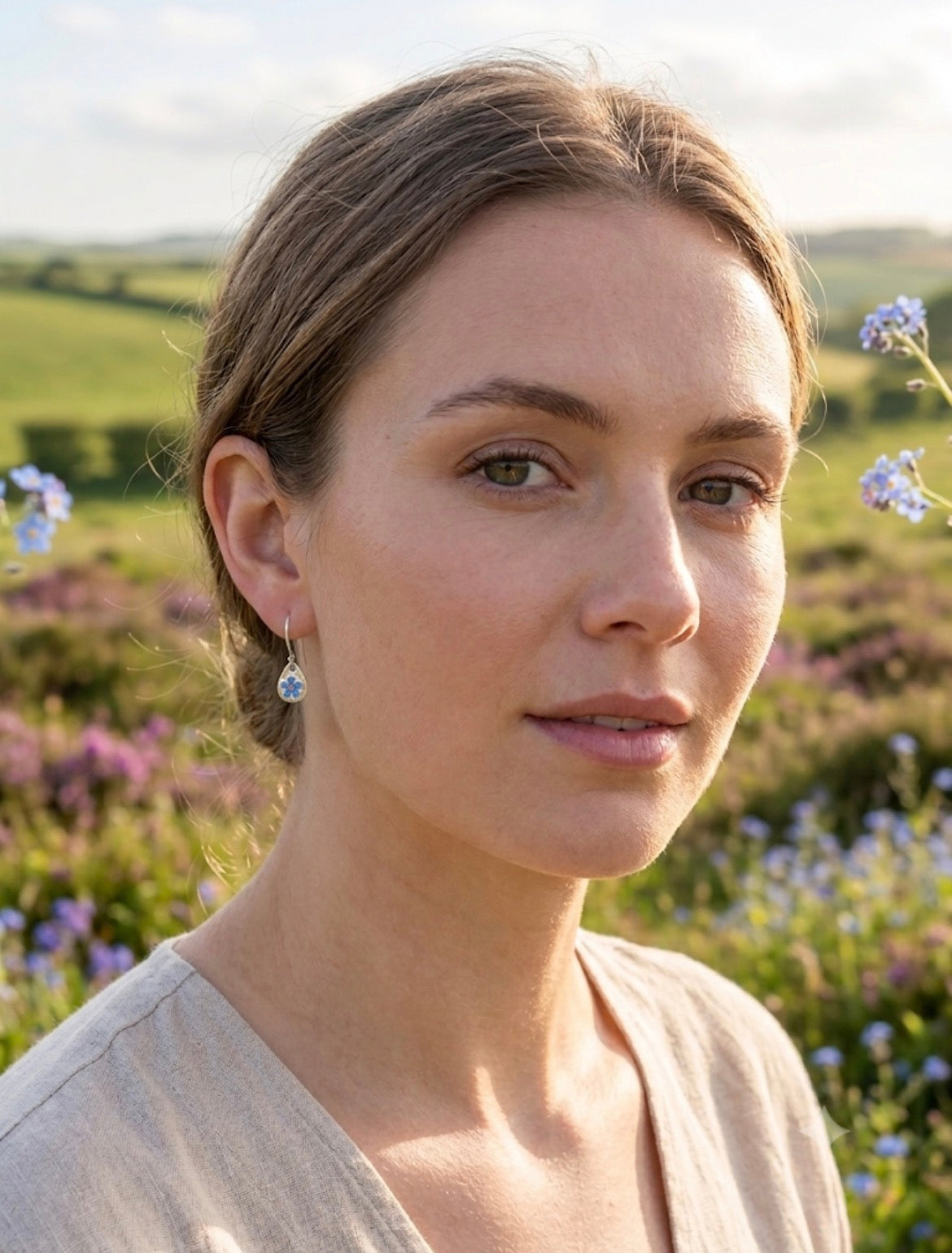 A woman, wearing small blue forget me not teardrop earrings, photographed outdoors in a field of purple flowers under a bright sky.
