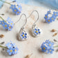 A pair of silver jewelery dangle earrings with small  forget me not flower charms, displayed on a textured beige fabric surrounded by small blue forget-me-not flowers.