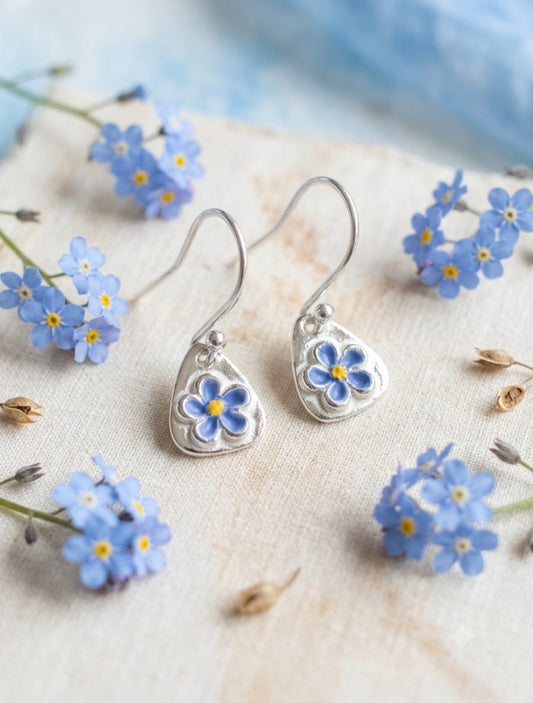 A pair of silver jewelery dangle earrings with small  forget me not flower charms, displayed on a textured beige fabric surrounded by small blue forget-me-not flowers.