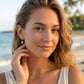 A woman wearing a small silver dangle whelk shell earring, posing on a sunny beach with the ocean and palm trees blurred in the background.