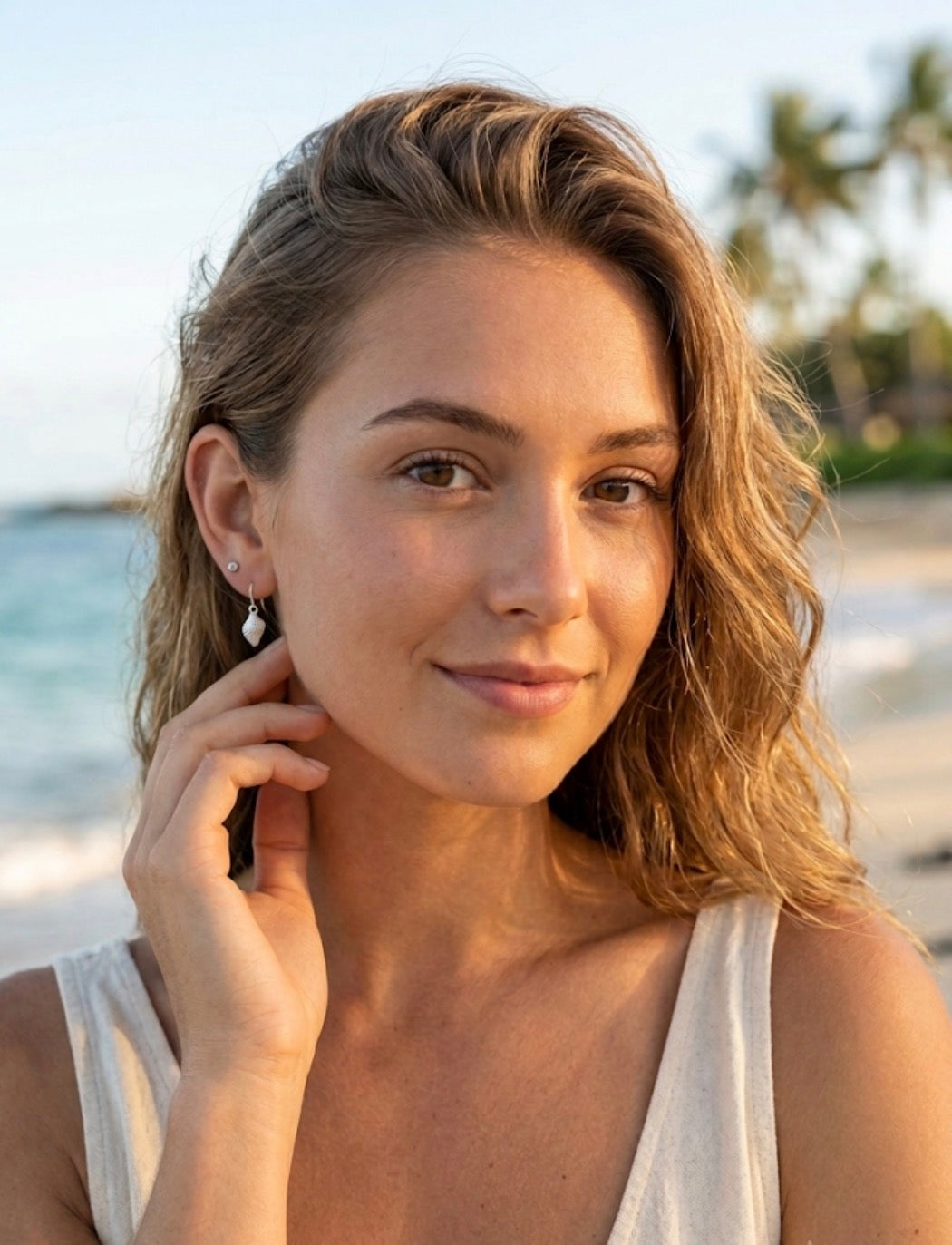 A woman wearing a small silver dangle whelk shell earring, posing on a sunny beach with the ocean and palm trees blurred in the background.