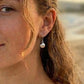 A close-up of a woman with light brown hair wearing a silver cockle shell dangle earring, set against a softly blurred beach background.