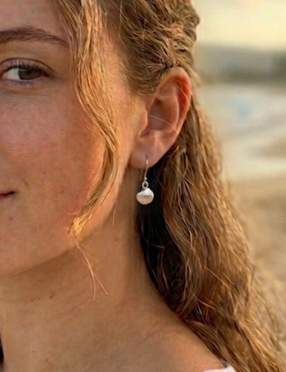 A close-up of a woman with light brown hair wearing a silver cockle shell dangle earring, set against a softly blurred beach background.