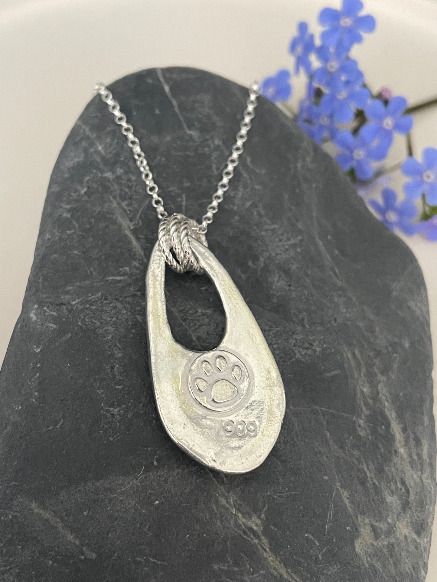 The reverse of the forget-me-not flower pendant showing the molly and me jewellery  paw print together with the 999 mark for fine silver on the pendant with a cutout and 3 jump rings attaching it to a silver chain. 