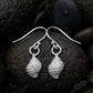 A pair of silver dangle earrings shaped like textured seashells, resting on dark wet river stones.