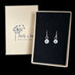 Pair of silver earrings with small sea urchins dangling from ear wires set in black velvet next to a Molly and Me Jewellery branded box. 