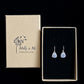 Teardrop shaped silver dangle earrings with hand painted forget me not flowers presented on black velvet next to its branded packaging box by Molly & Me Jewellery.