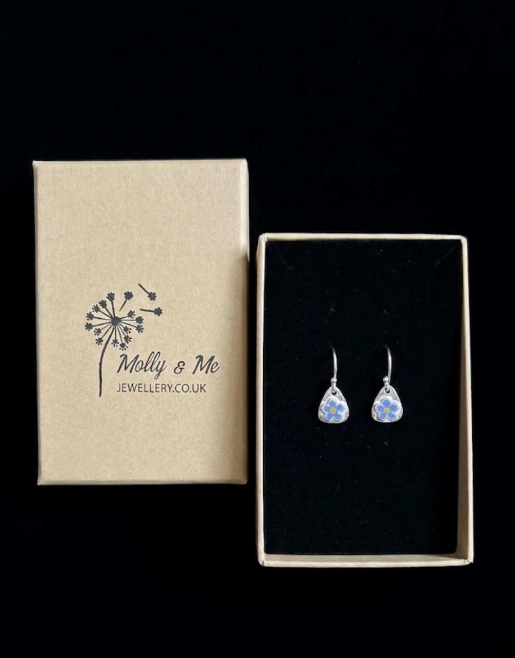 Teardrop shaped silver dangle earrings with hand painted forget me not flowers presented on black velvet next to its branded packaging box by Molly & Me Jewellery.