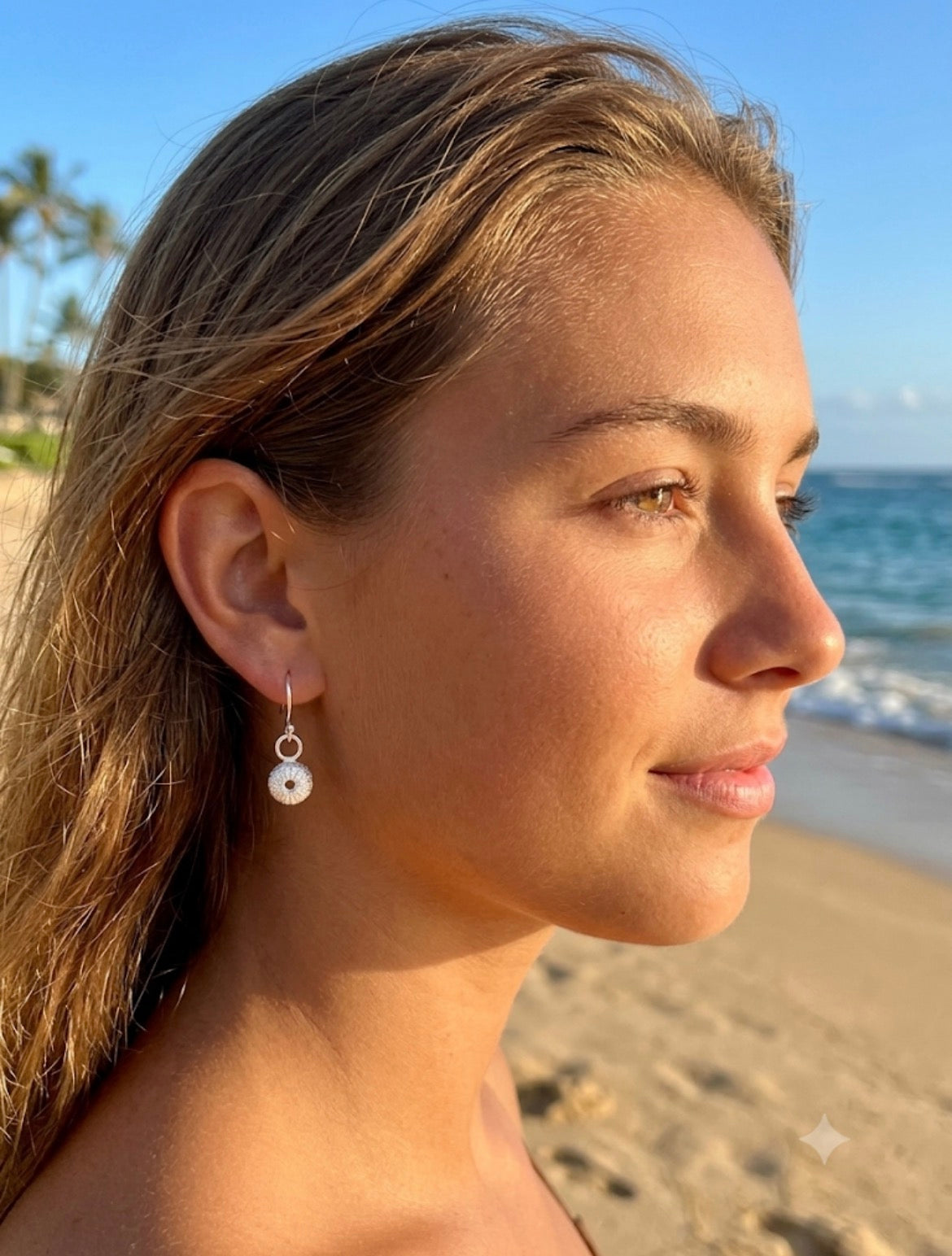 A woman in profile at a beach wearing a delicate silver drop earring with a sea urchin charm. 
