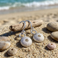 Silver sea urchin dangle earrings displayed on a sandy beach with driftwood and seashells, with the ocean waves blurred in the background.