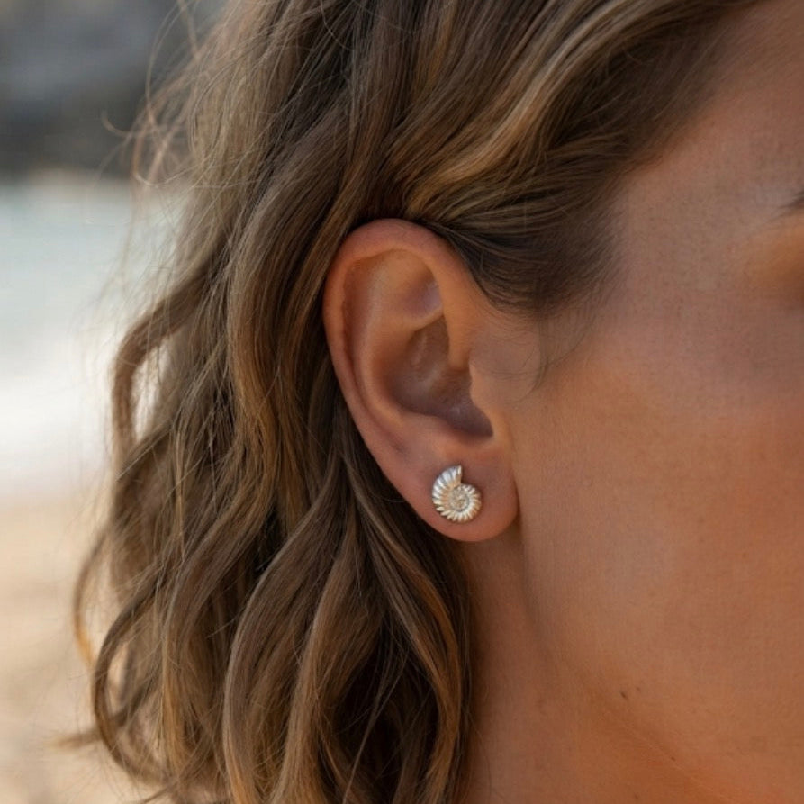 A close-up of a woman's ear wearing a small silver ammonite fossil shell earring with a set against a blurred beach background.