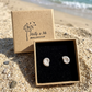 A pair of silver spiral shell ammonite stud earrings in an open black-lined jewelry box on sandy beach ground, with the branded Molly and Me Jewellery kraft box lid standing behind them and the ocean in the background.