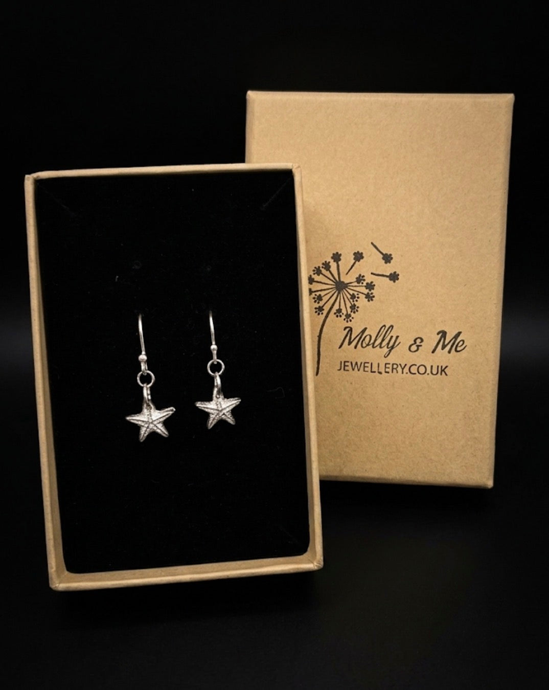 Silver starfish dangle earrings with detailed surface displayed in an open black-lined gift box next to its kraft paper packaging box branded 'Molly & Me Jewellery’. 