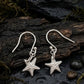 A pair of fine silver starfish dangle earrings displayed on a dark, wet piece of wood with small stones and moss around them.