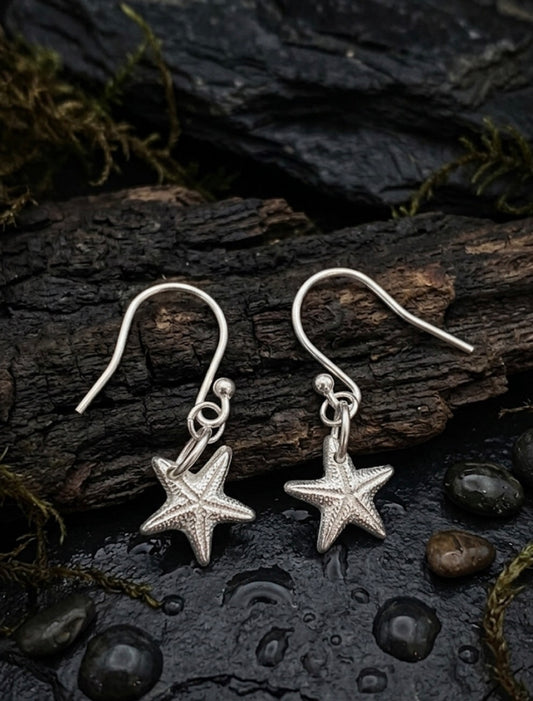 A pair of fine silver starfish dangle earrings displayed on a dark, wet piece of wood with small stones and moss around them.
