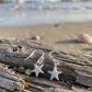 A pair of silver starfish dangle earrings resting on a piece of weathered driftwood on a sandy beach, with the ocean waves blurred in the background.