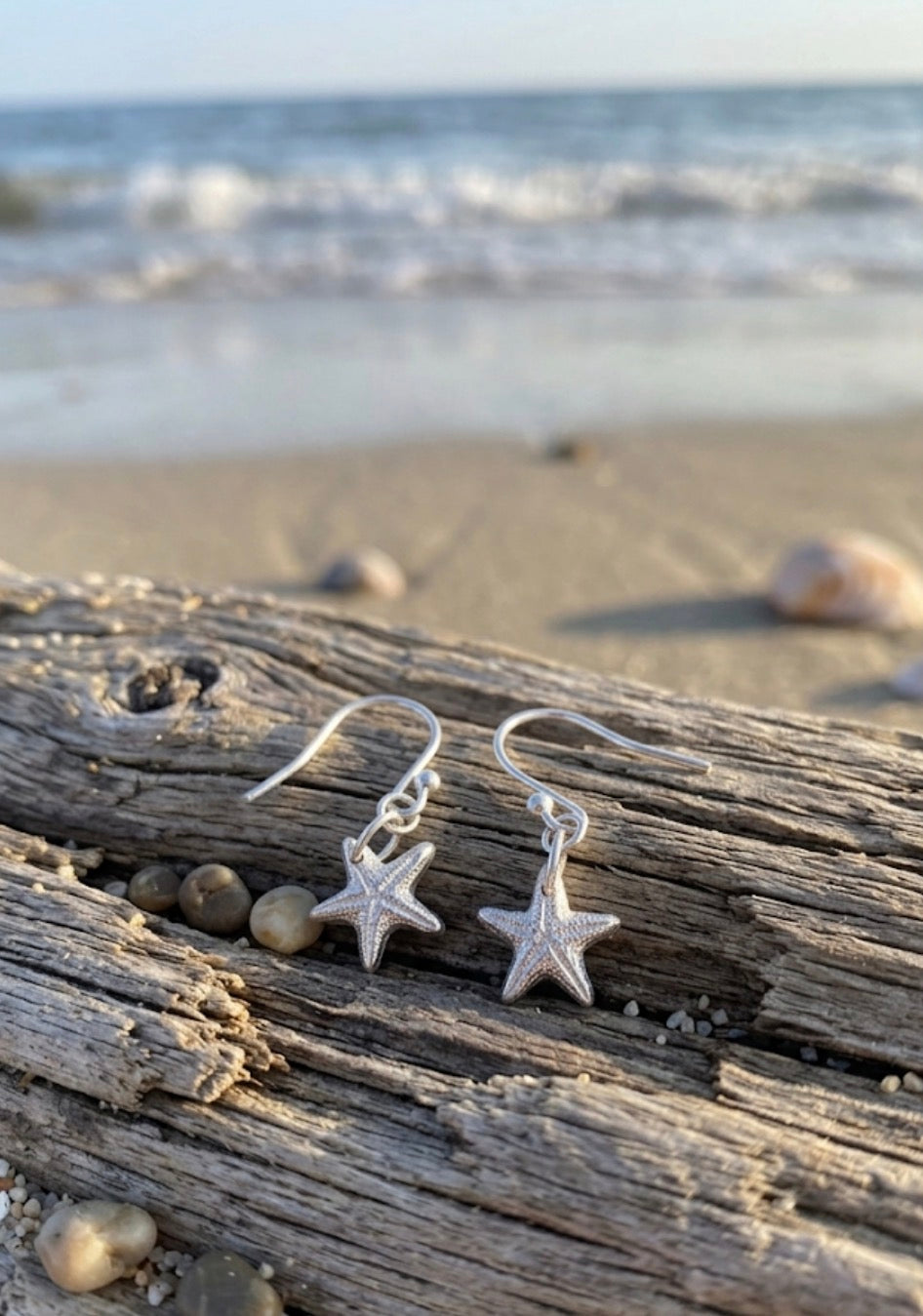 A pair of silver starfish dangle earrings resting on a piece of weathered driftwood on a sandy beach, with the ocean waves blurred in the background.