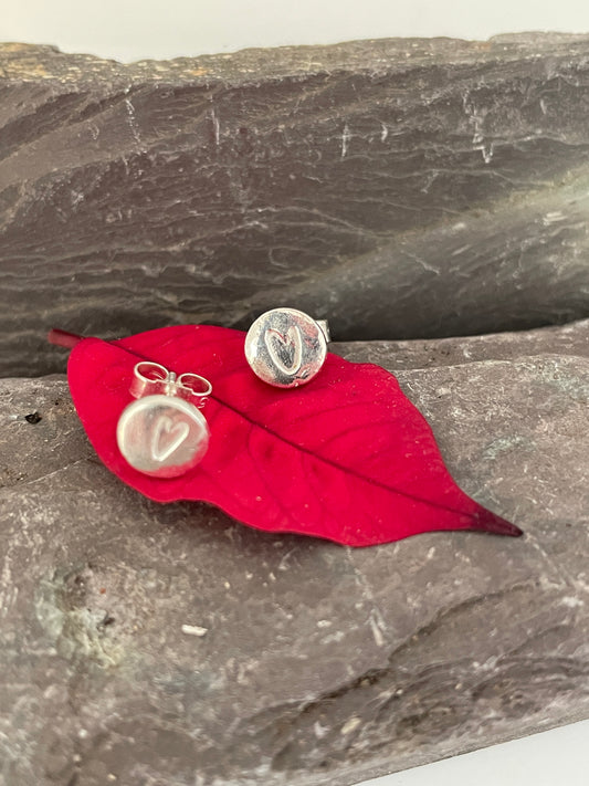 Shiny disc stud earrings with an impression of a tiny love heart in the middle, set on a red leaf on a stone