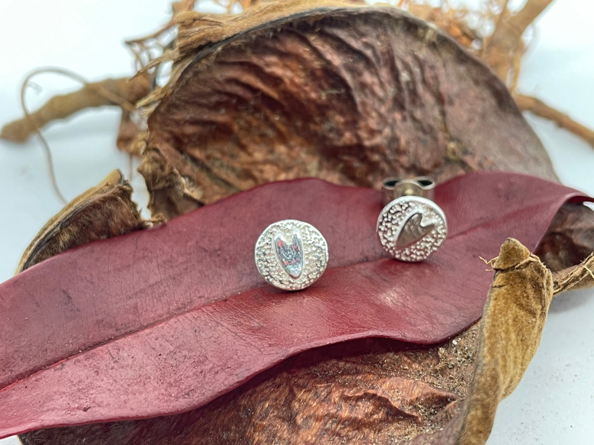 Tiny textured disc shaped earrings with smooth tiny love hearts on them set on a natural background.