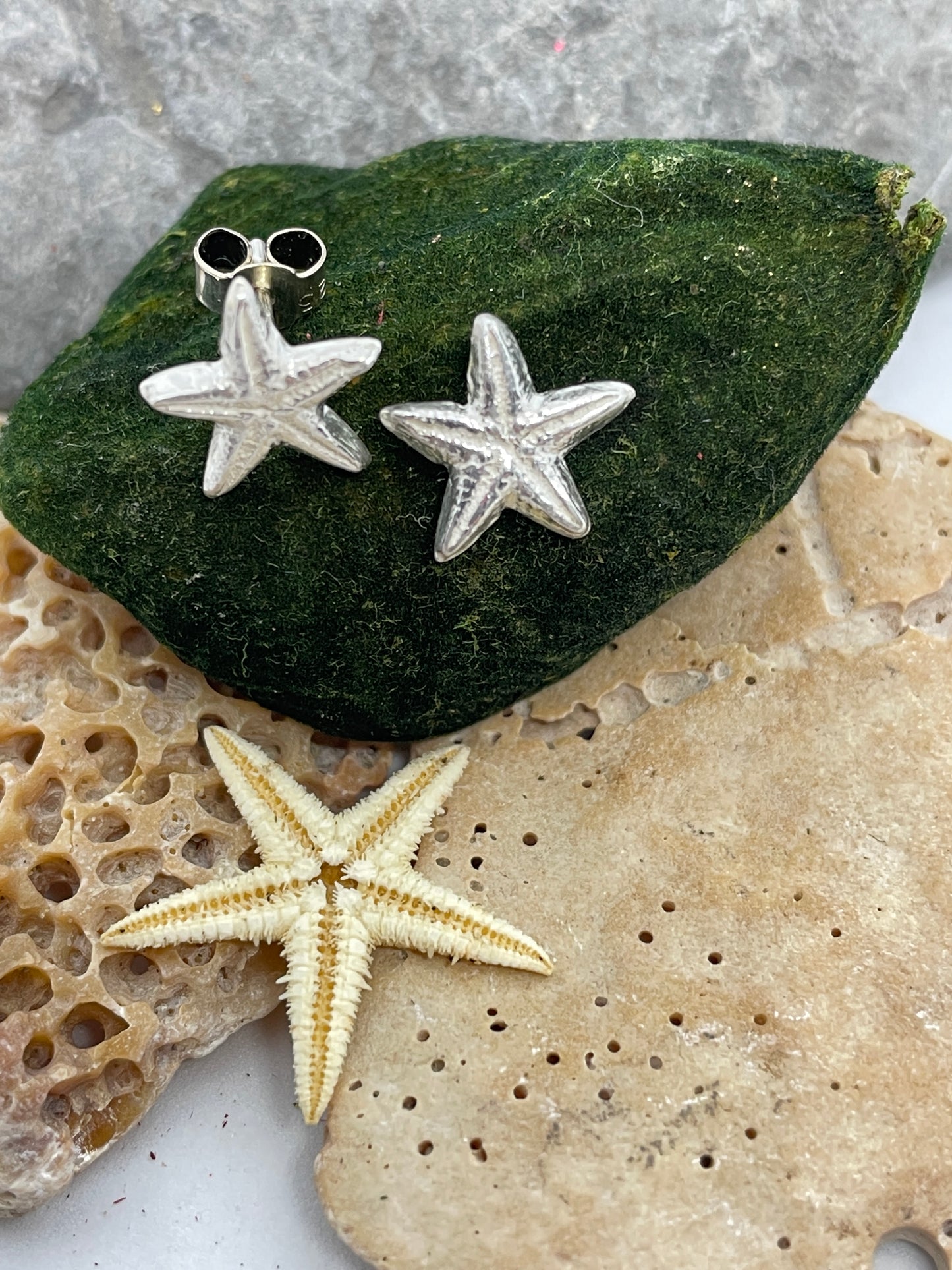 Small silver starfish stud earrings set on a green moss background with pebbles and a small real starfish in the foreground